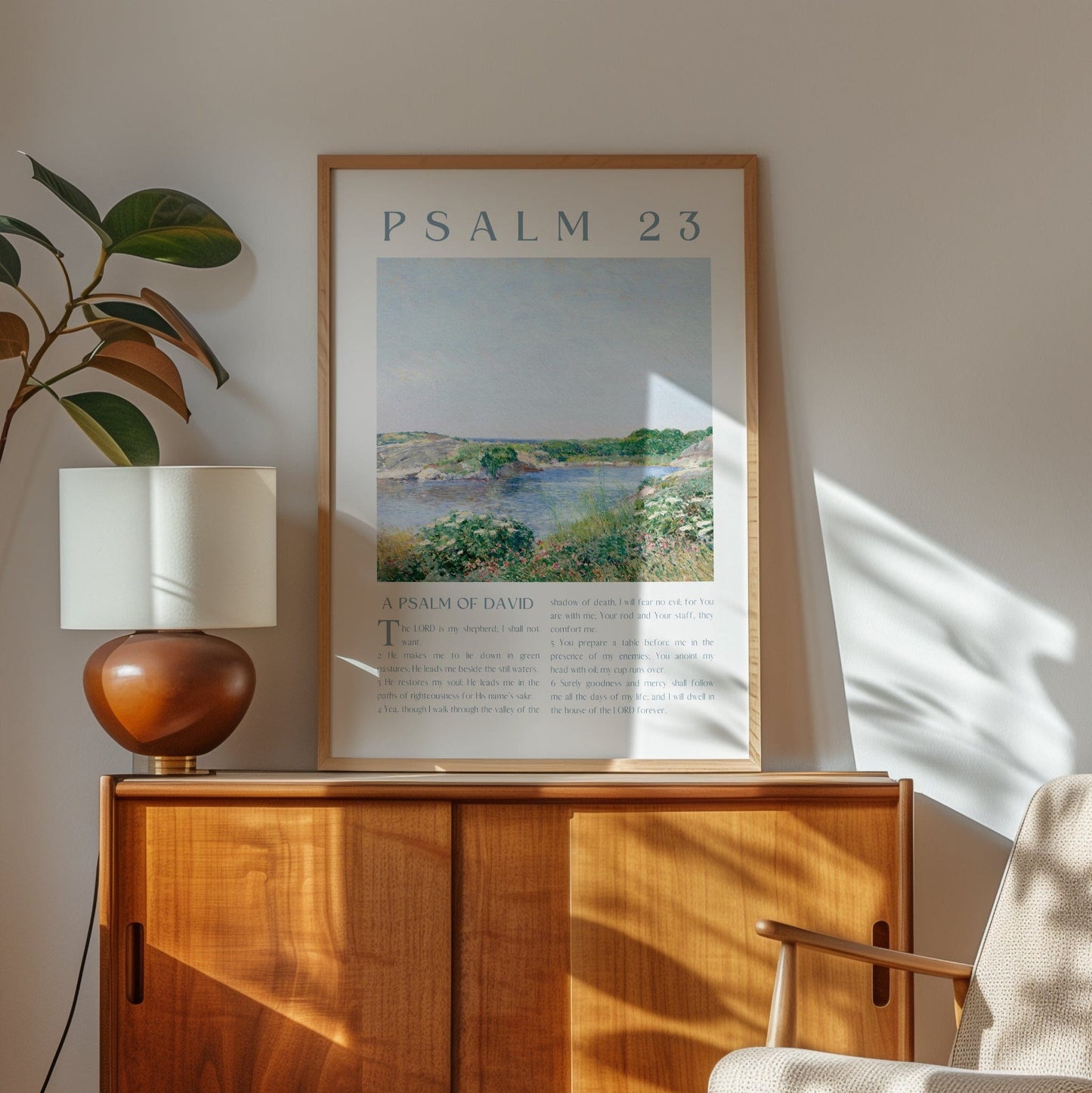 Framed artwork titled 'Psalm 23' on a wooden cabinet with a lamp and plant in a room.