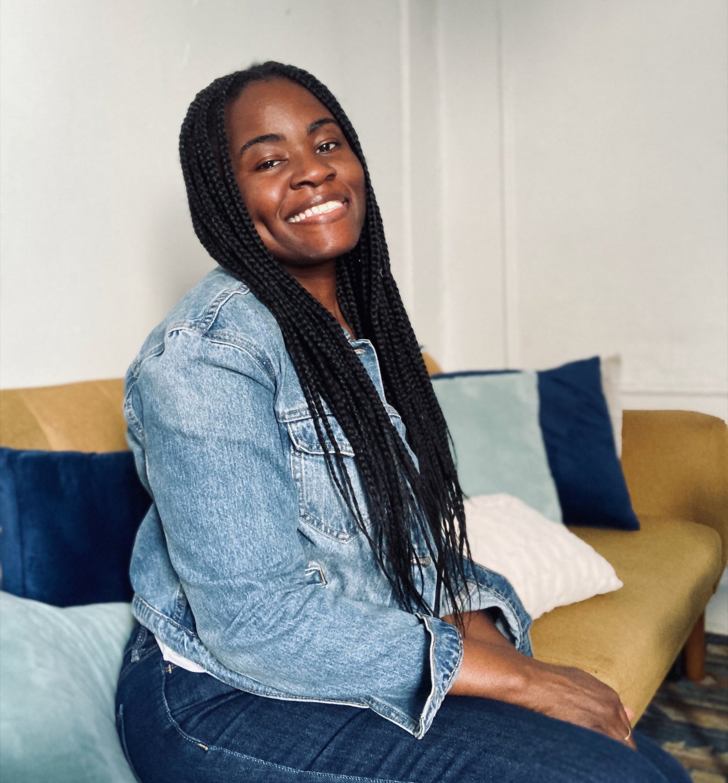 Woman sitting on a couch wearing a denim jacket and jeans, smiling.