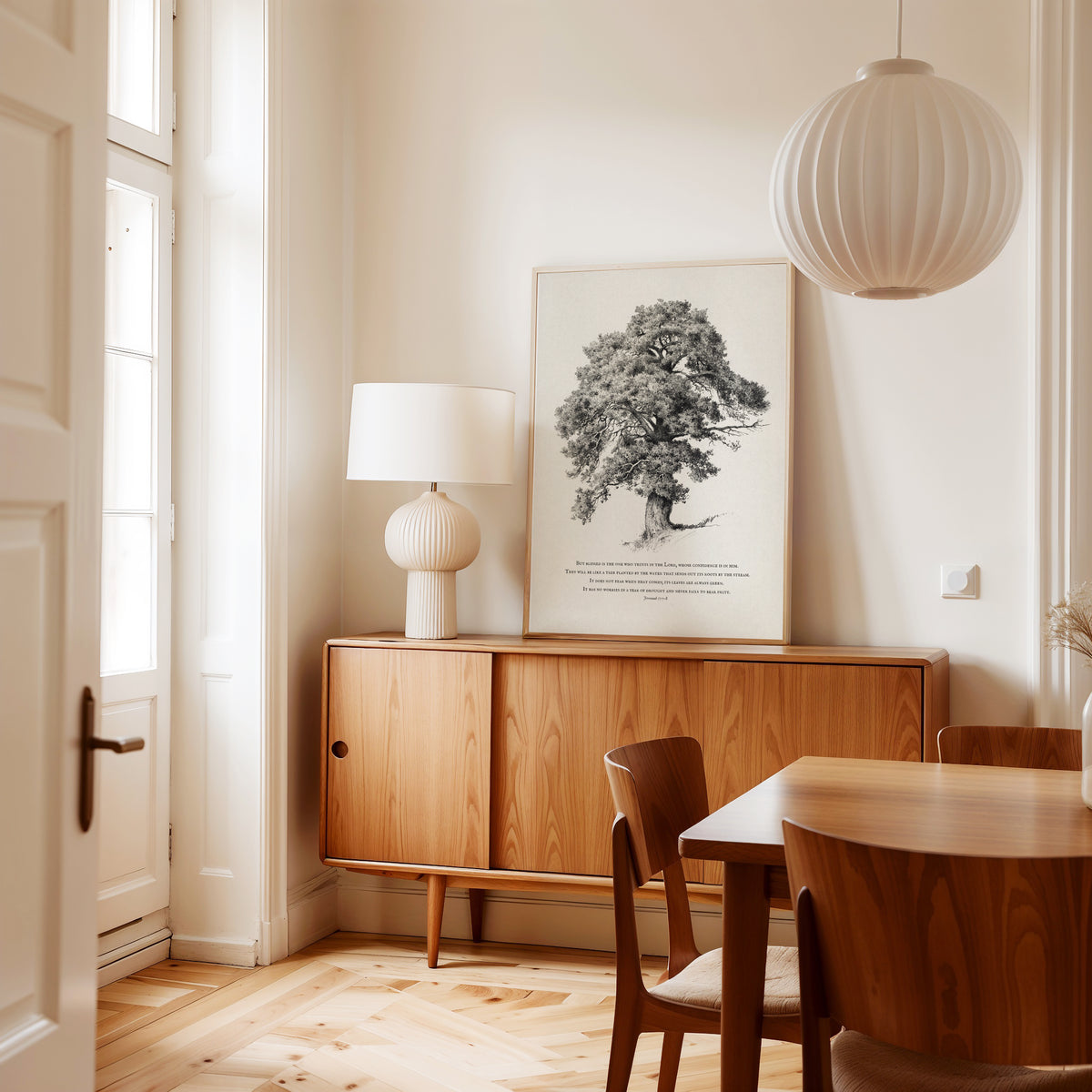 A styled interior corner featuring a mid-century wooden credenza with a white table lamp, a large black-and-white framed tree print on the wall, a white pleated pendant light, and a wooden dining table with chairs in a warm, sunlit room.