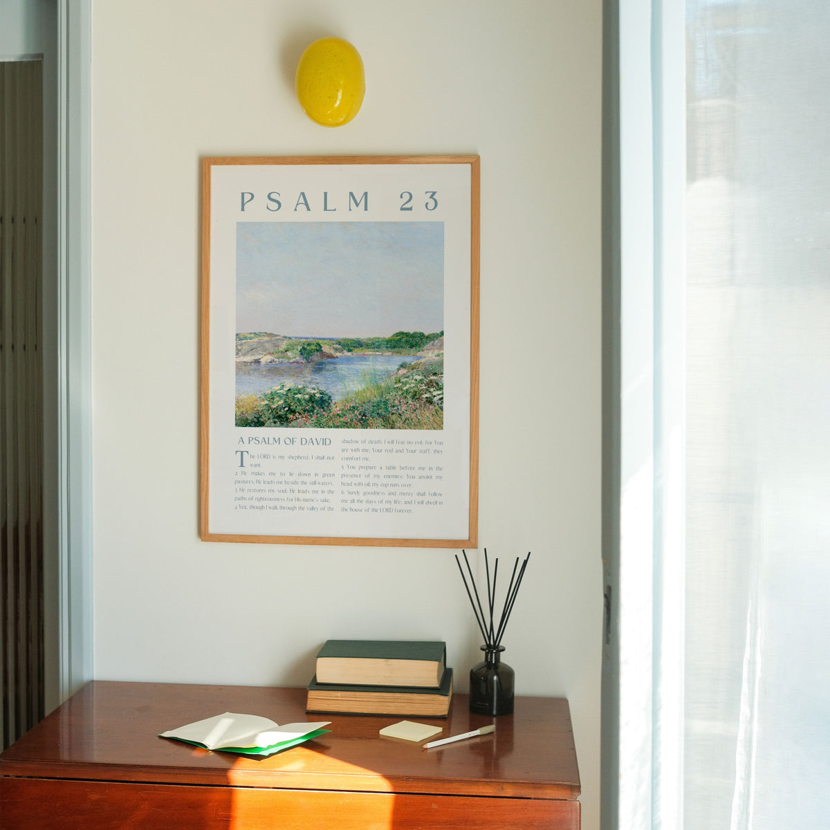 A framed art print of Psalm 23 with a landscape painting and text, hanging on a light beige wall above a wooden console table with books, a reed diffuser, and an open notebook. A yellow wall lamp is visible above the frame.