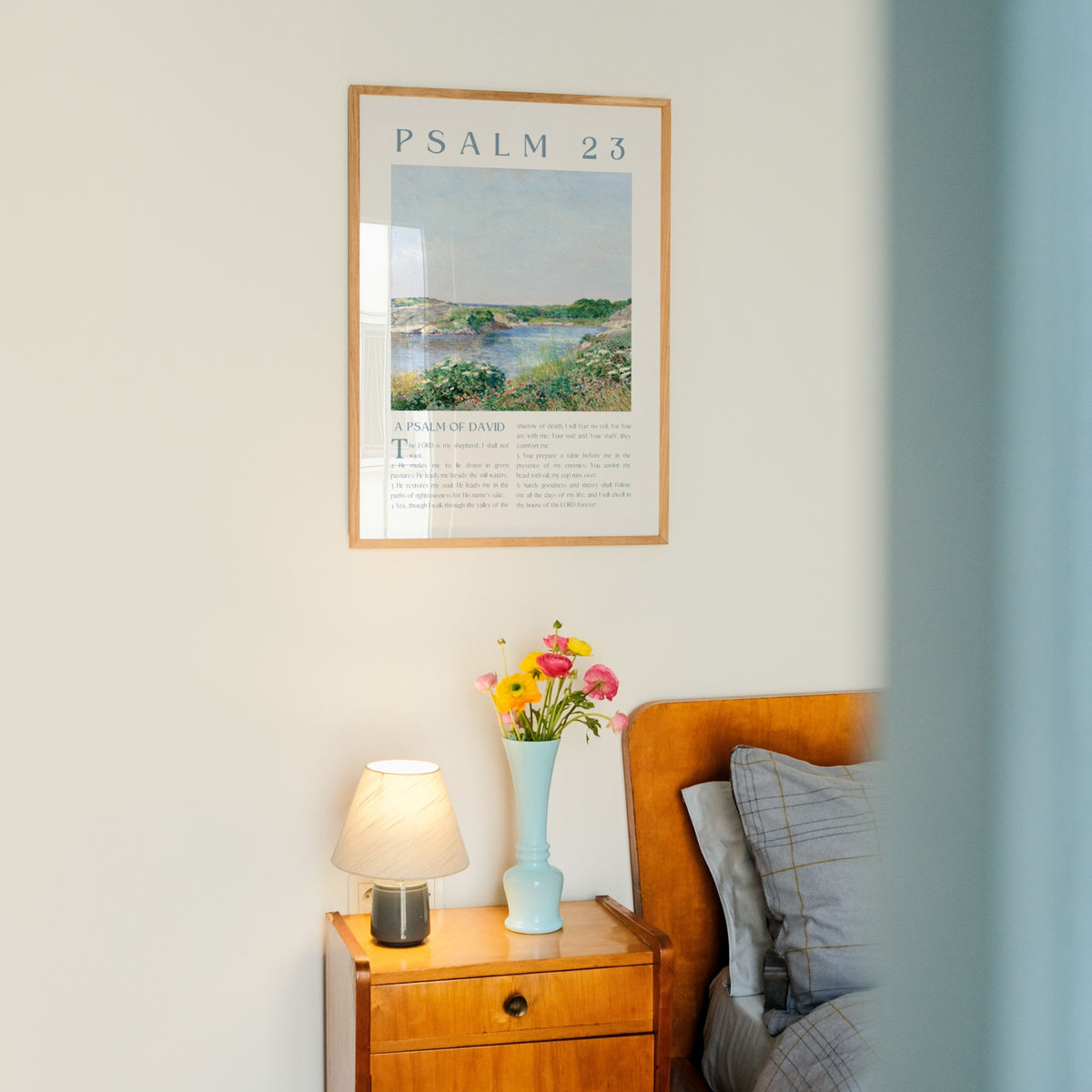 A framed art print of Psalm 23 hanging on a light-colored wall above a wooden bedside table with a lamp and a vase of flowers.