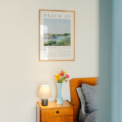 A framed art print of Psalm 23 hanging on a light-colored wall above a wooden bedside table with a lamp and a vase of flowers.