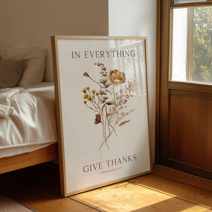 Framed artwork with floral design and text 'In everything give thanks' on a wall in a room with a bed and window.