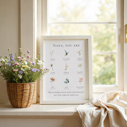 Framed print with floral illustrations and text on a windowsill with flowers and a blanket.
