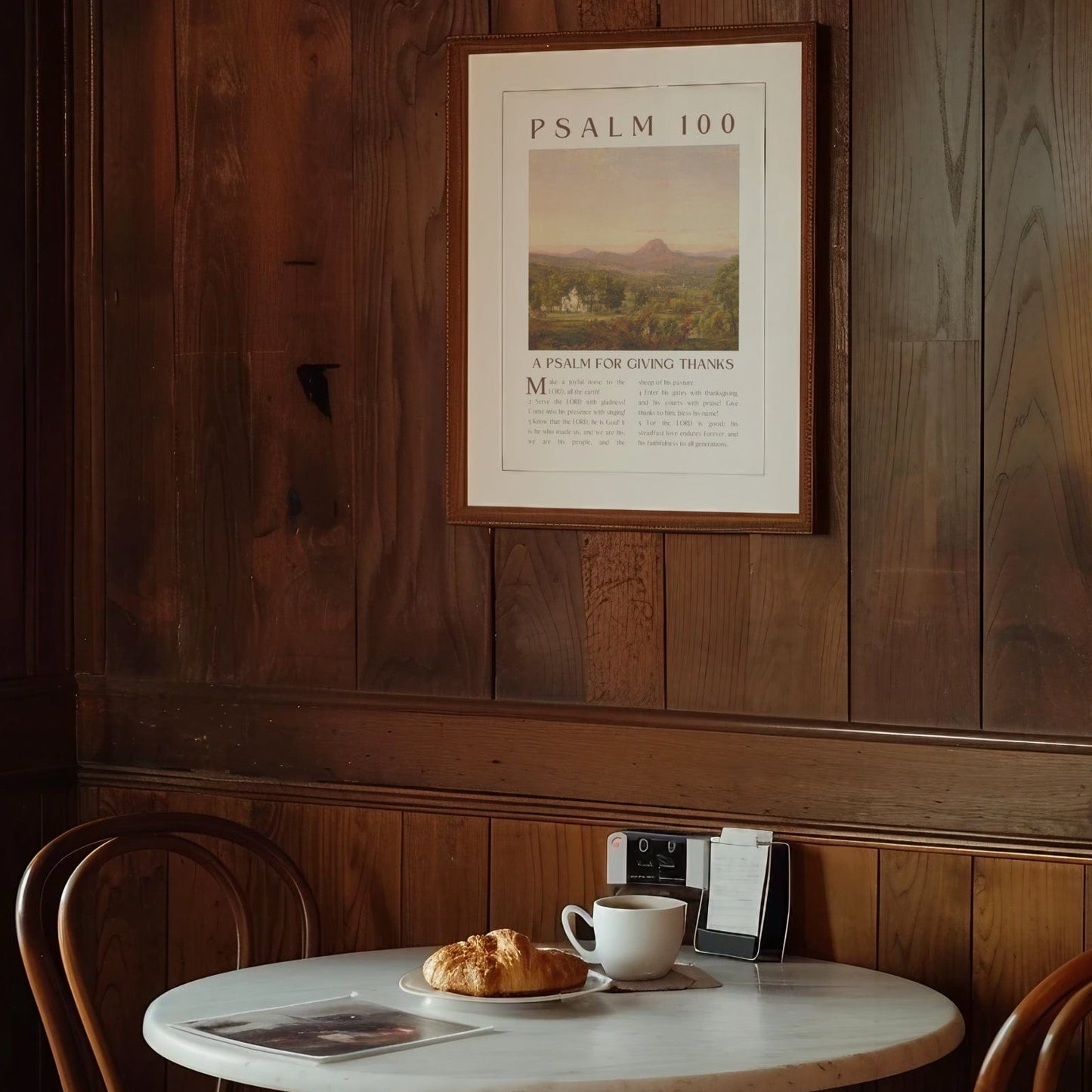 Café interior with a table set for two, a cup of coffee, and a croissant, framed poster on the wall.