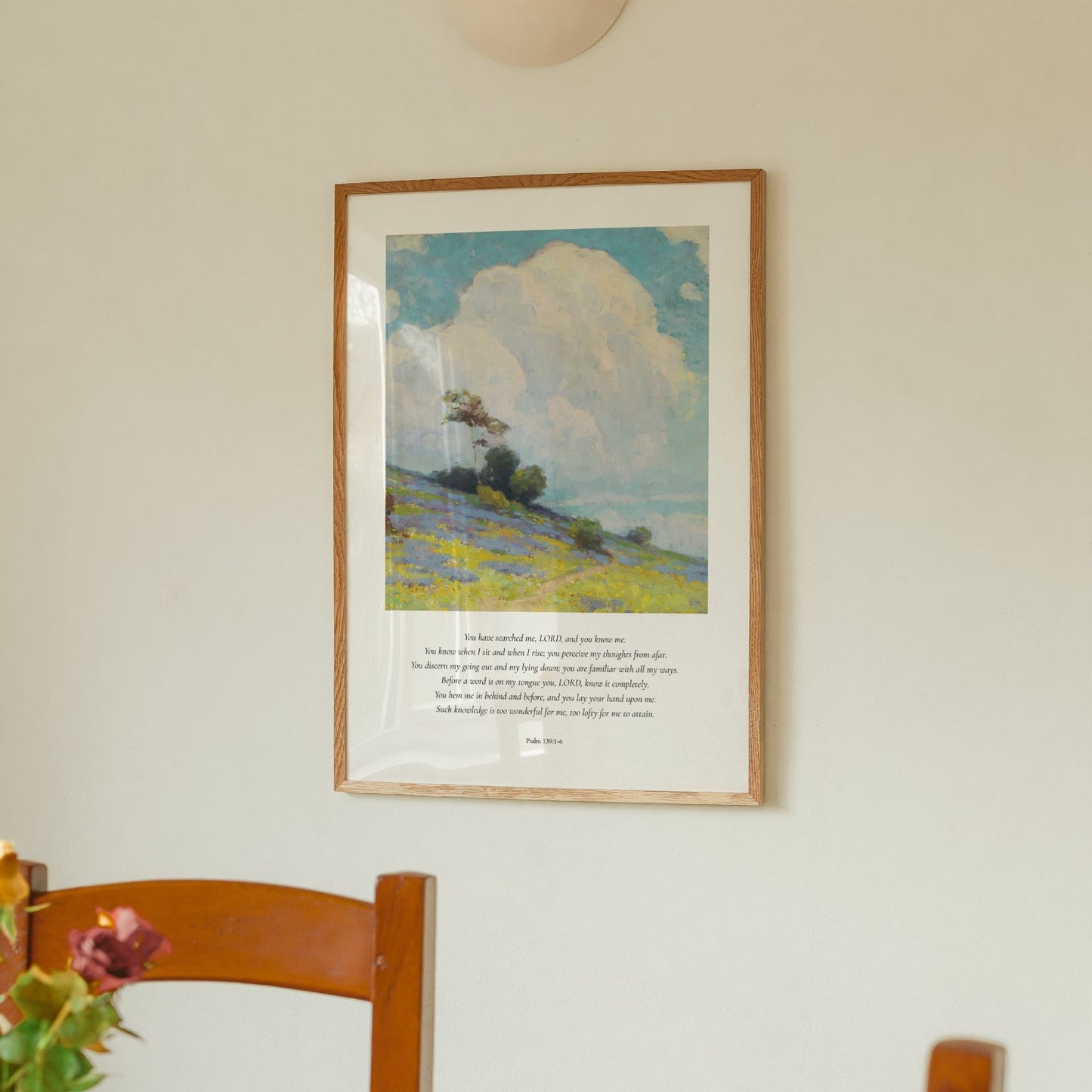 Framed landscape painting on a wall above a wooden chair with flowers.