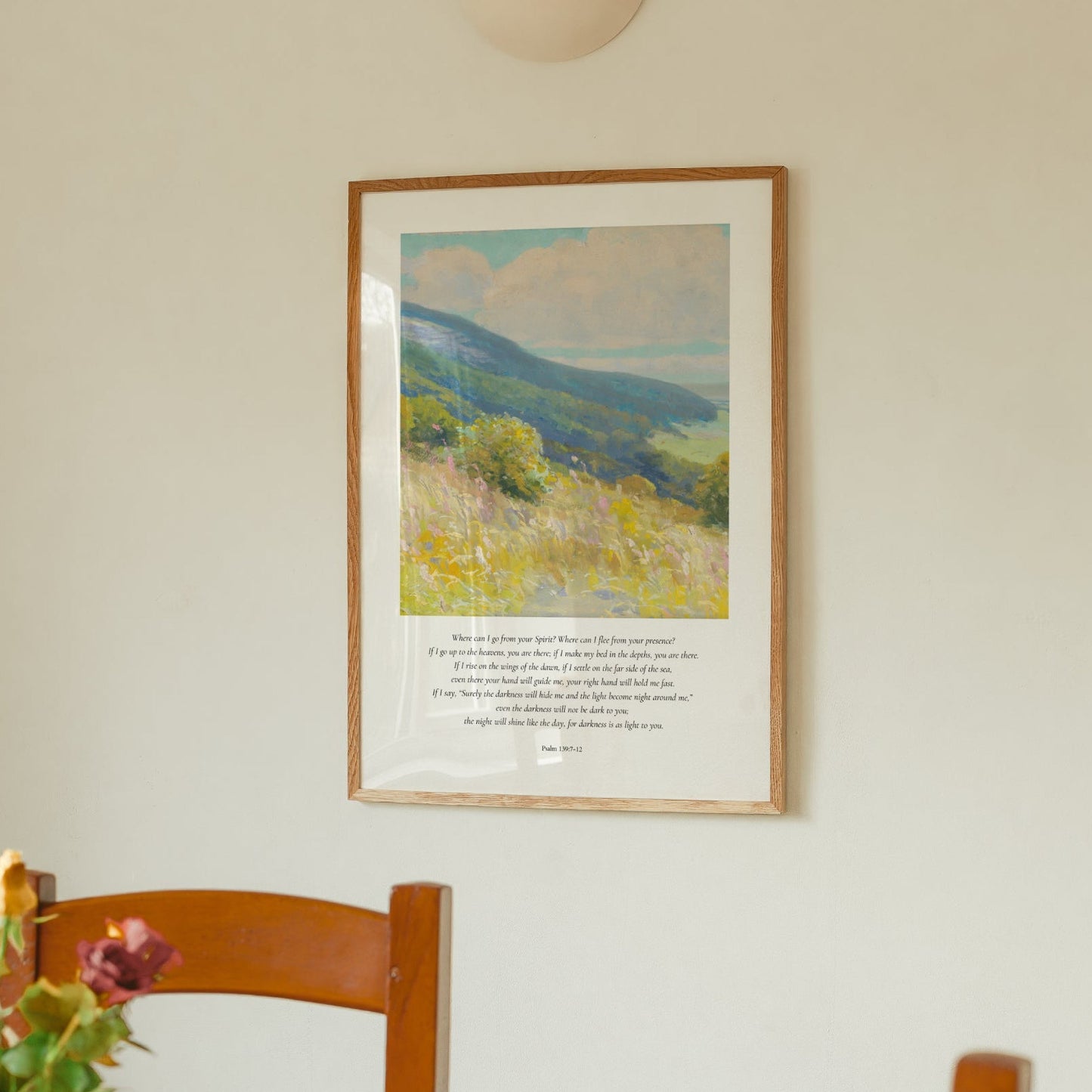 Framed landscape painting on a wall above a wooden chair with flowers.