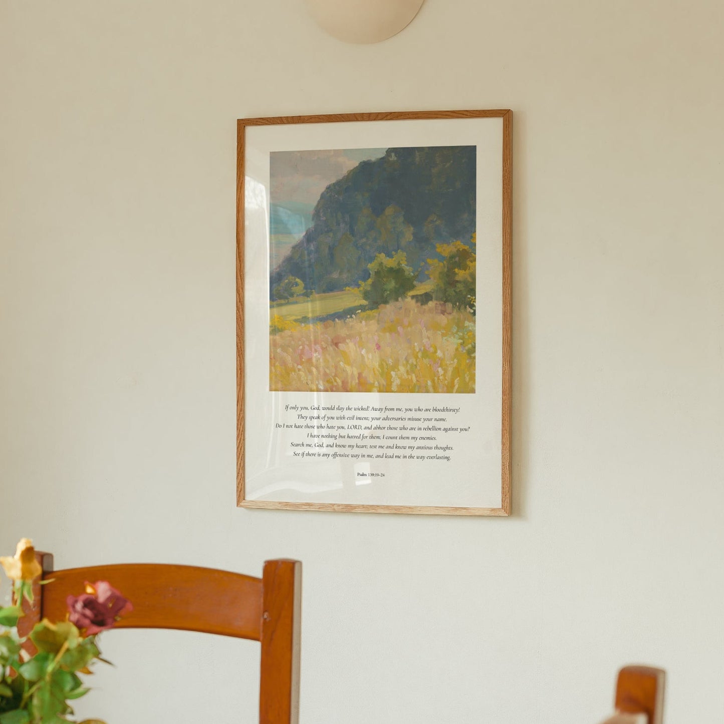 Framed landscape print on a wall above a wooden chair with flowers.