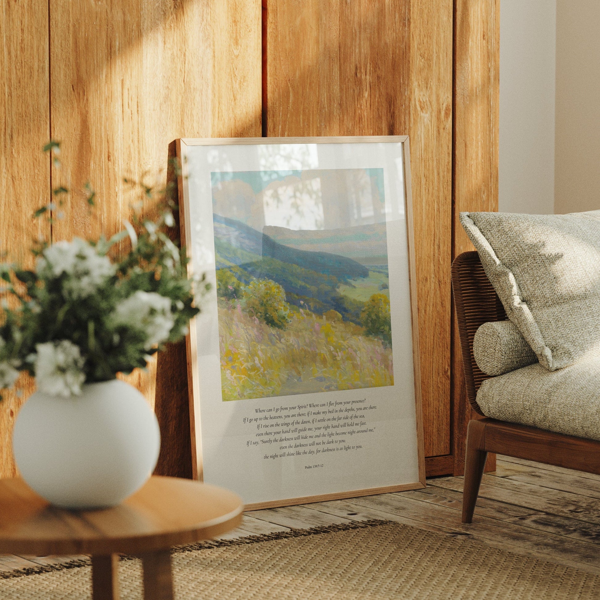 Framed landscape print on a wooden wall with a vase of flowers and a chair in the foreground.