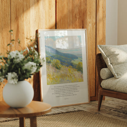 Framed landscape print on a wooden wall with a vase of flowers and a chair in the foreground.