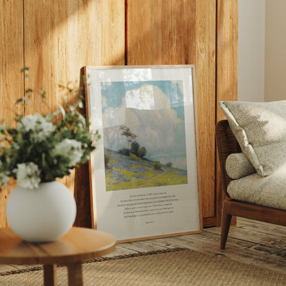 Framed landscape print on a wooden wall with a vase of flowers and a chair in the foreground.