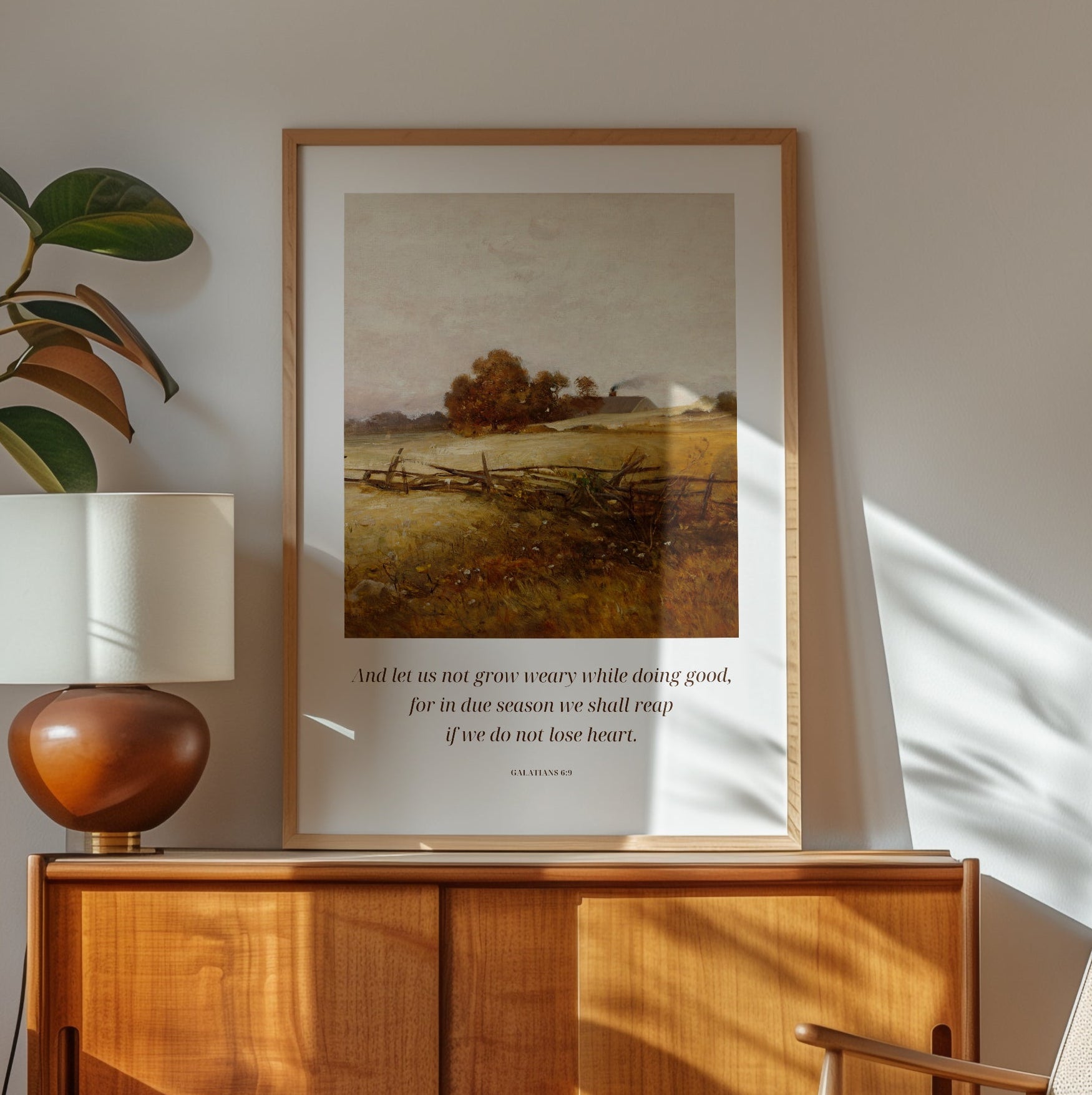 Framed landscape print on a wall above a wooden cabinet with a lamp and chair.